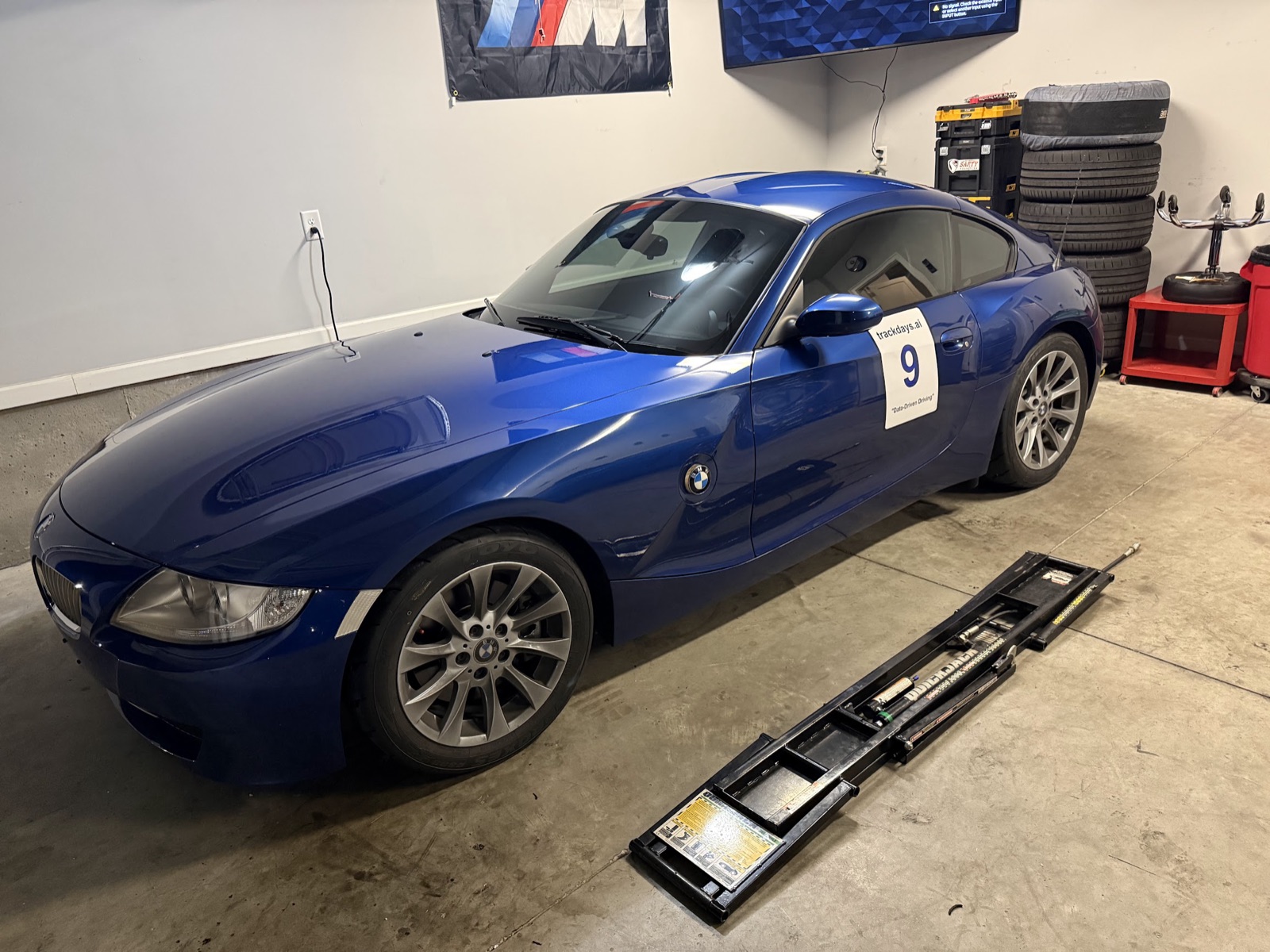 The 2008 Z4 3.0si Coupe in Bill's garage, Montego Blue, wearing the number 9 trackdays.ai door magnet, still on stock wheels and Toyo tires, with a stack of spare tires and a jack ramp beside it